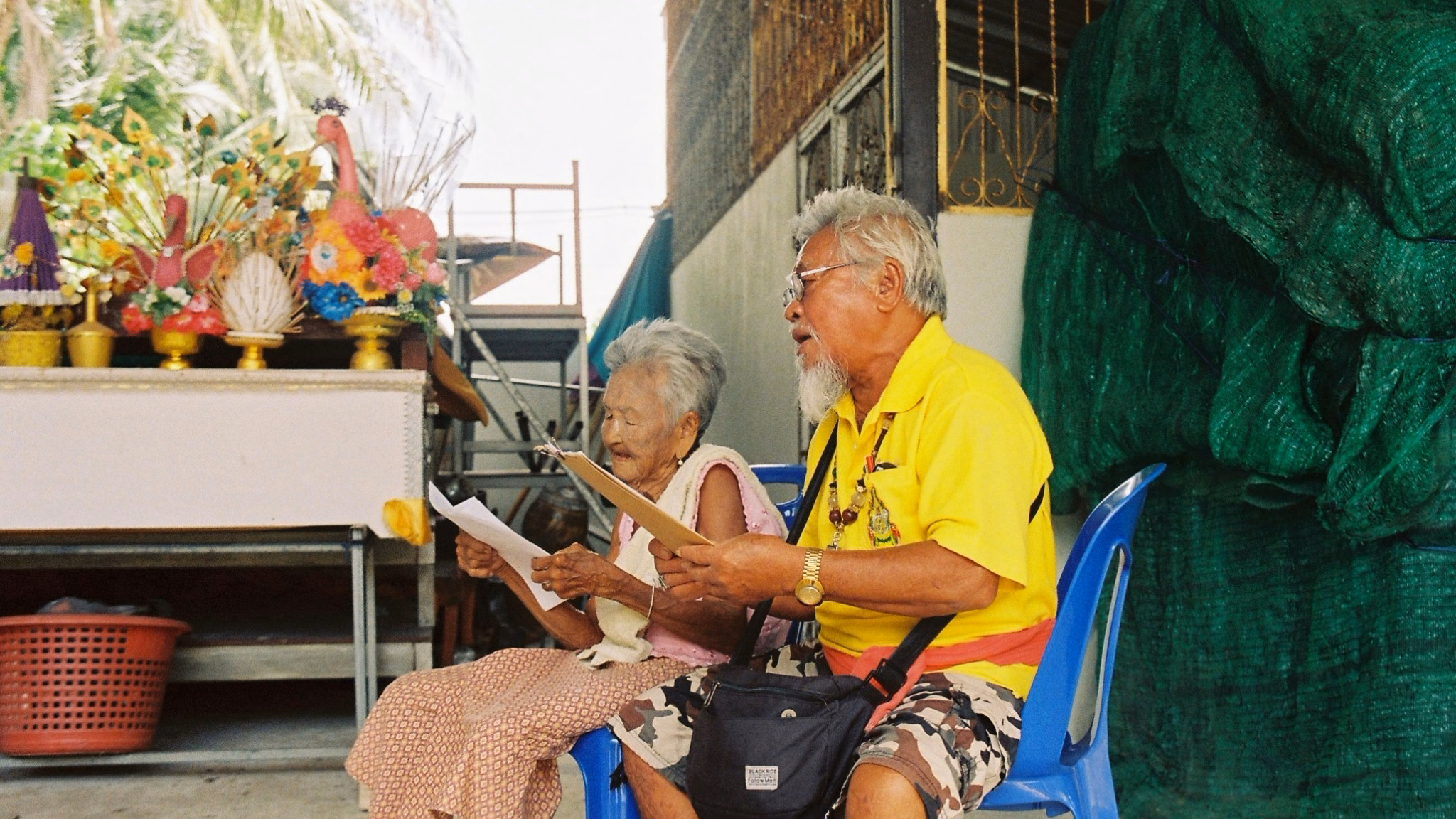 ผู้สูงอายุชาวเกาะพะงันสองคนกำลังขับร้องเพลงกาพย์เห่เรือ นั่งอยู่หน้าศาลาที่ประดับดอกไม้